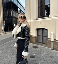 Frau in schwarzer Leder-Teddyjacke und Sonnenbrille auf städtischer Straße.