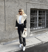 Frau in schwarzer Leder-Teddyjacke, weißem Crop-Top, Jeans und Sneakers vor Betonwand.