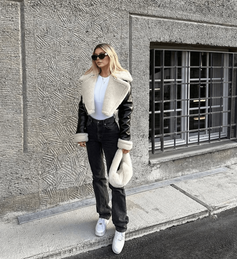 Frau in schwarzer Leder-Teddyjacke, weißem Crop-Top, Jeans und Sneakers vor Betonwand.