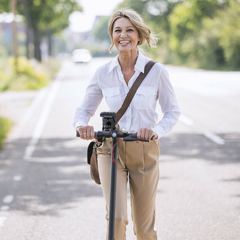 Frau auf E-Scooter, weiße Bluse, beige Hose, sonniger Tag, Stadtstraße, umweltfreundliche Mobilität.
