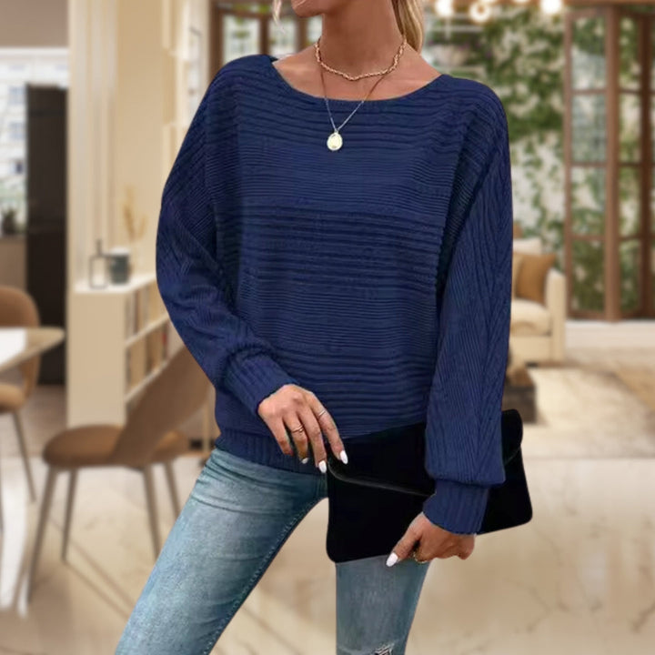 Frau in blauem Strickpullover und Jeans, hält schwarze Clutch, modernes Wohnzimmer im Hintergrund.