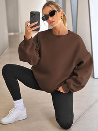Frau in braunem Oversize-Pullover, schwarzen Leggings, Sonnenbrille, kniet, Smartphone in Hand.