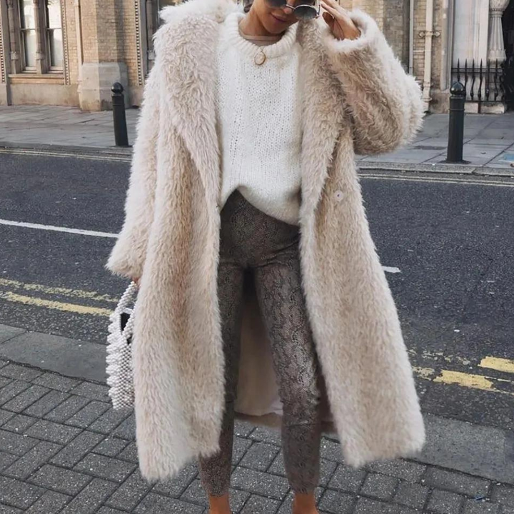 Frau in beigem Kunstpelzmantel, weißem Pullover und gemusterten Hosen auf städtischer Straße.