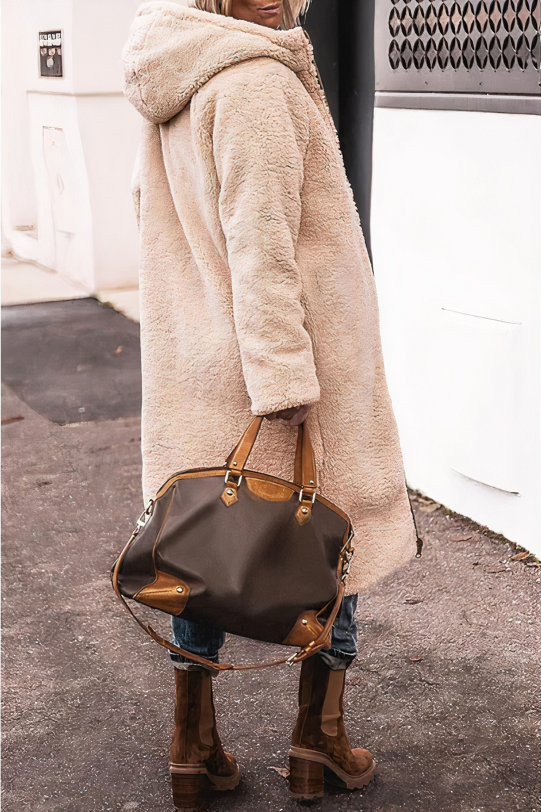 Frau in beigem Teddy-Mantel mit brauner Lederhandtasche und Stiefeln auf Gehweg.