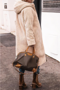 Frau in beigem Teddy-Mantel mit brauner Lederhandtasche und Stiefeln auf Gehweg.