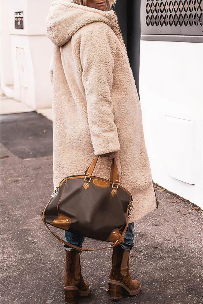Frau in beigem Teddy-Mantel mit brauner Lederhandtasche und Stiefeln auf Gehweg.