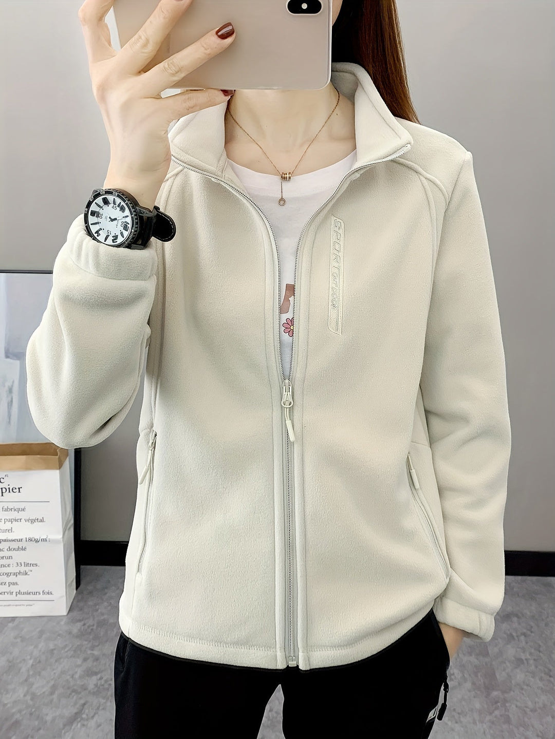 Frau in weißer Fleecejacke mit Reißverschluss, Smartphone, Armbanduhr, Innenraum.