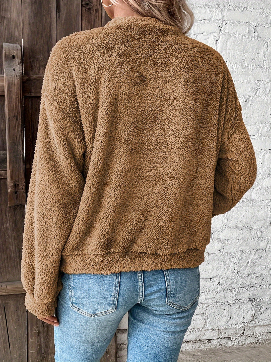 Frau in brauner Teddyjacke und blauer Jeans vor weißer Backsteinwand.