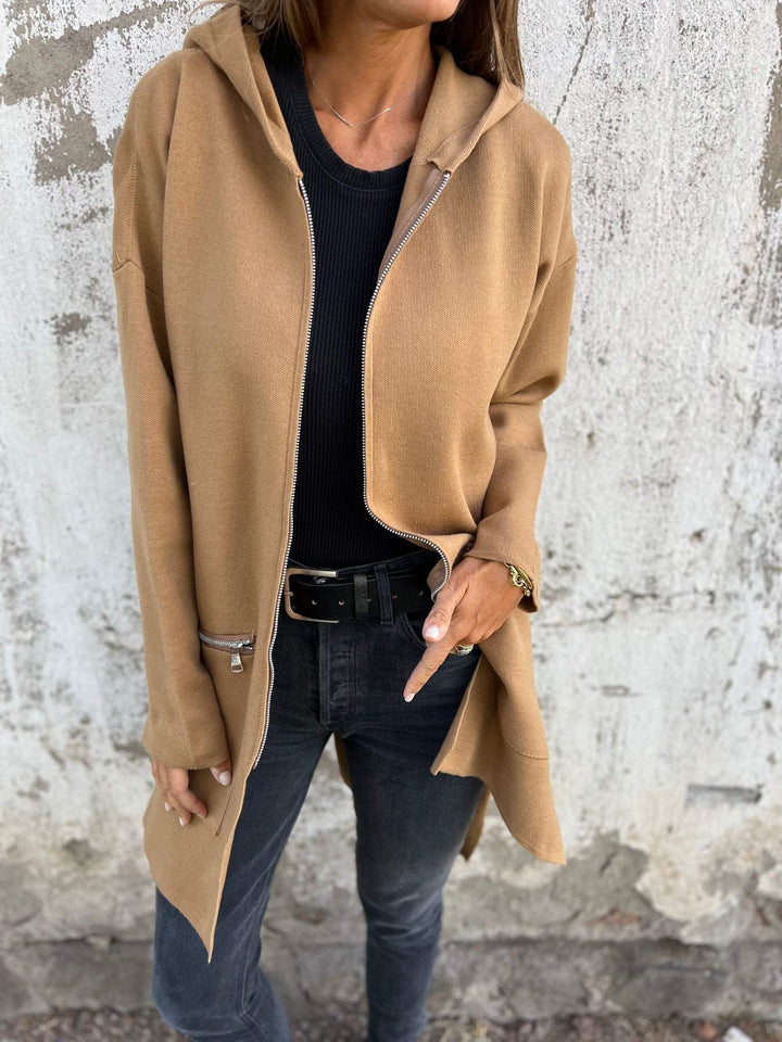 Frau trägt beige Kapuzenjacke mit Reißverschluss, schwarze Jeans, lässiger Look.