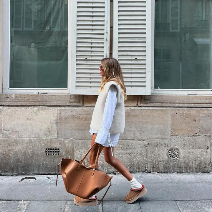 Frau mit brauner Ledertasche, weißem Kleid und Weste, modisch, urbaner Hintergrund.