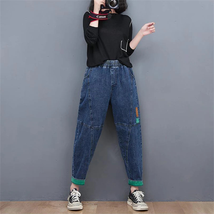 Frau in schwarzem Pullover und blauer Baggy-Jeans, lässiger Stil, grauer Hintergrund.