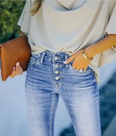 Frau in blauer Jeans mit Knopfleiste und beiger Bluse, hält braune Lederhandtasche.