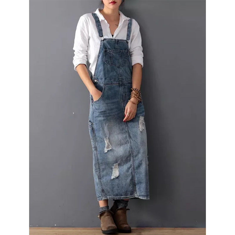 Frau in zerrissenem Jeans-Latzkleid, weißer Bluse, braunen Stiefeln vor grauer Wand.