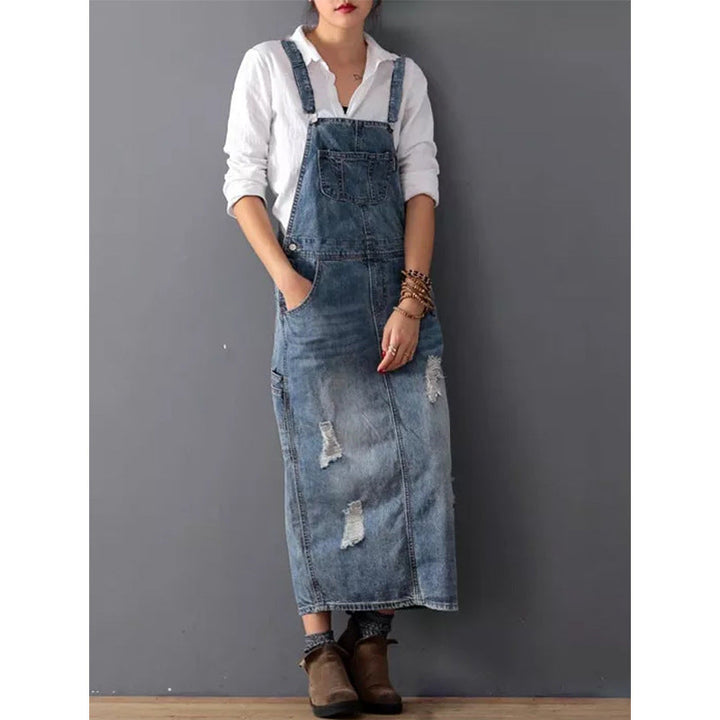 Frau in zerrissenem Jeans-Latzkleid, weißer Bluse, braunen Stiefeln vor grauer Wand.