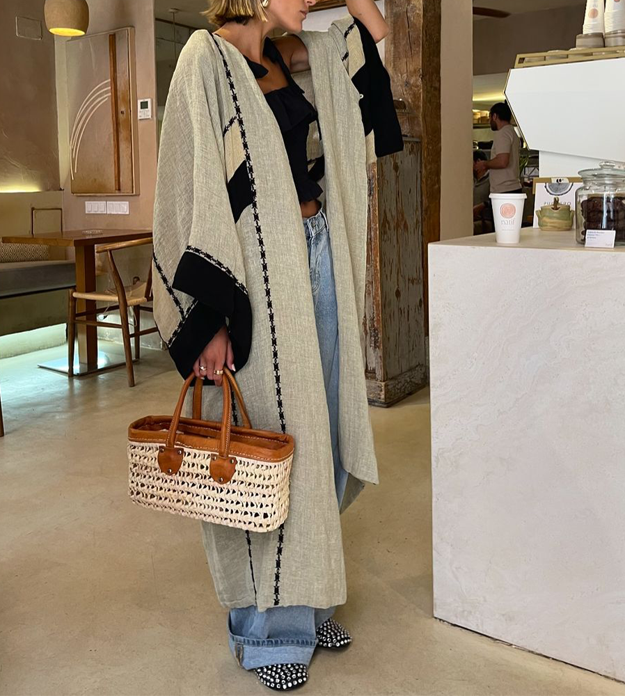 Frau in beigem Kimono mit schwarzem Muster, Jeans und Korbtasche in modernem Café.