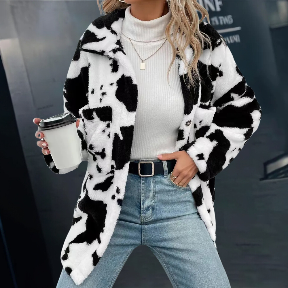 Frau in schwarz-weißem Kuhfellmantel, weißem Rollkragenpullover, Jeans, hält Kaffeebecher.