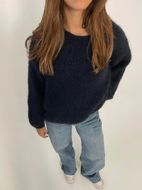 Frau in dunkelblauem Strickpullover und blauer Jeans, weiße Sneaker, lässiger Look.