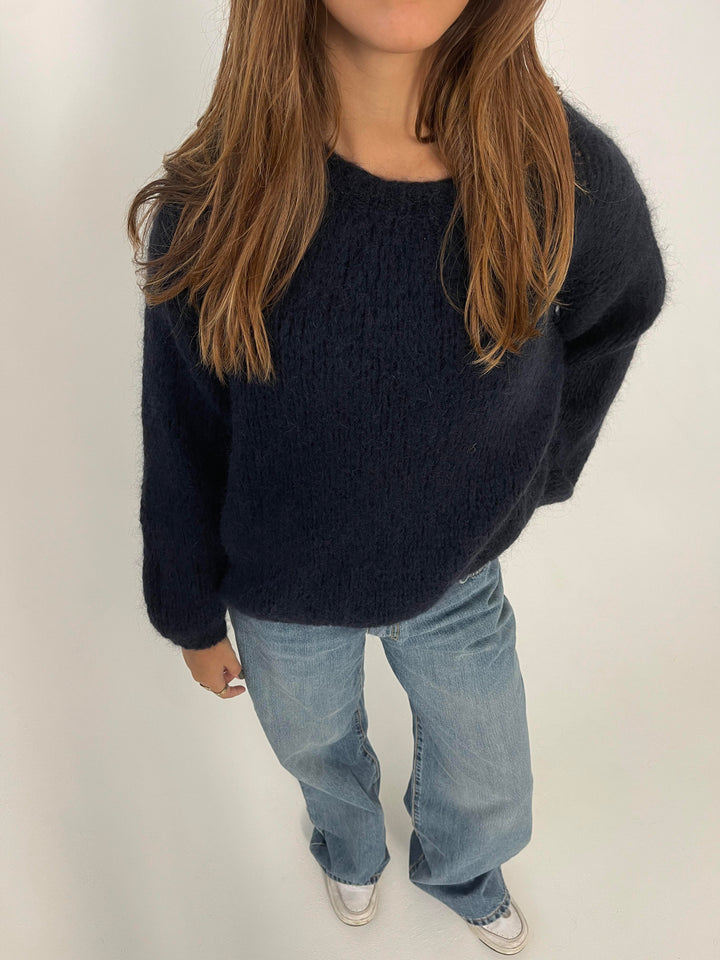 Frau in dunkelblauem Strickpullover und blauer Jeans, weiße Sneaker, lässiger Look.