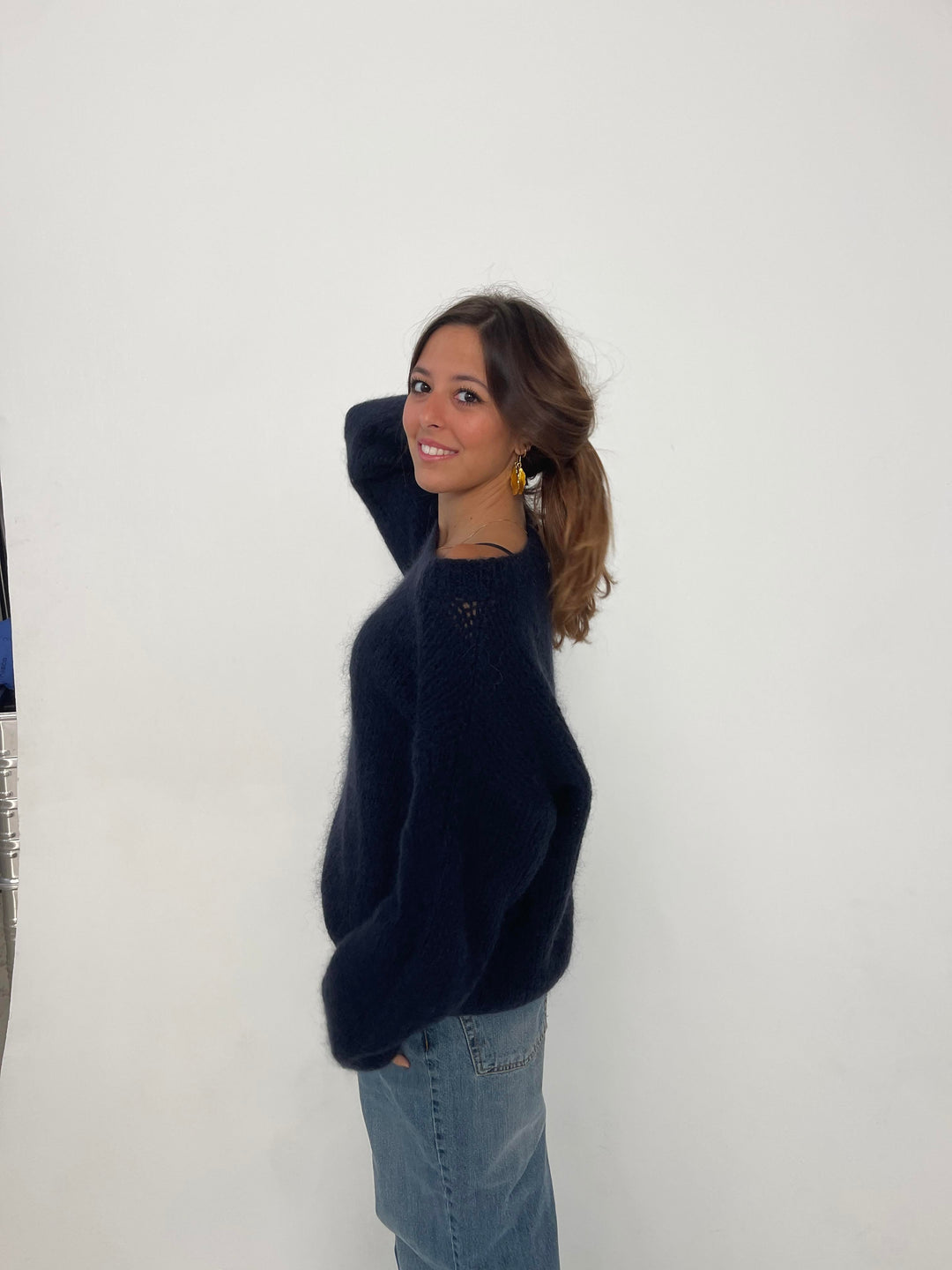 Frau in dunkelblauem Pullover und Jeans vor weißem Hintergrund, lächelnd, seitlich stehend.