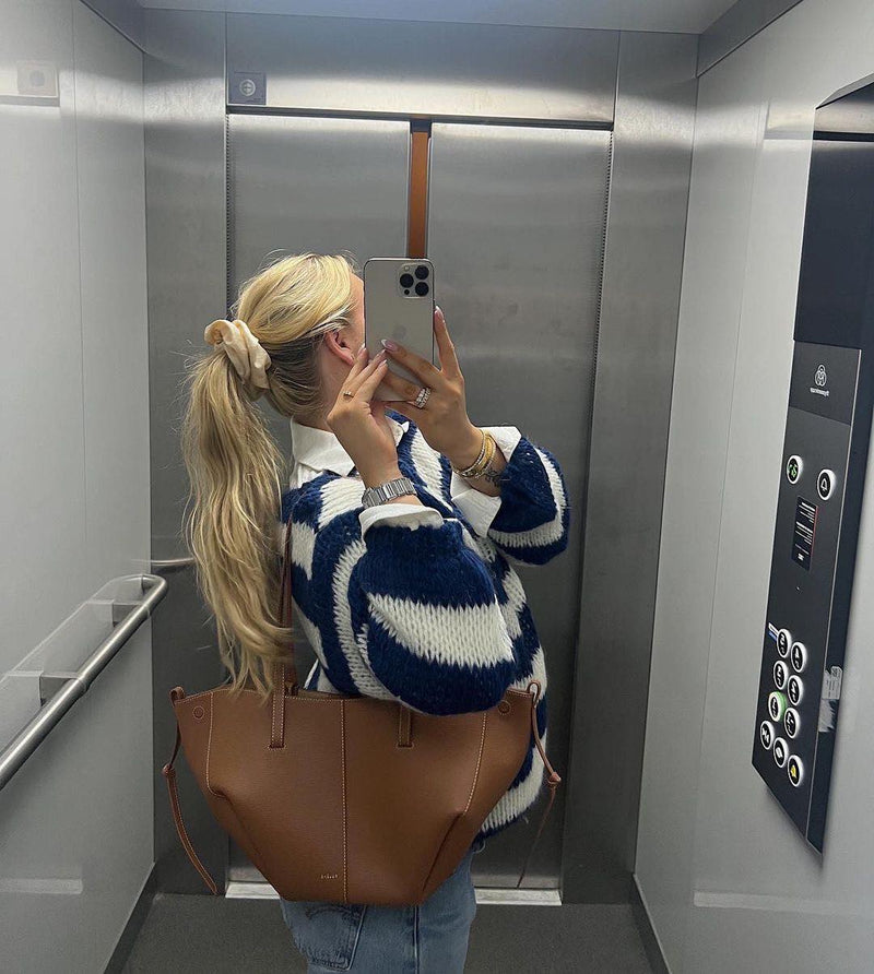 Frau im Aufzug mit blau-weißem Pullover, brauner Tasche, iPhone, blonden Haaren, Haargummi.