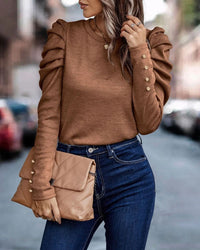 Frau in braunem Pullover mit Puffärmeln, blaue Jeans, beige Clutch, Stadtstraße im Hintergrund.