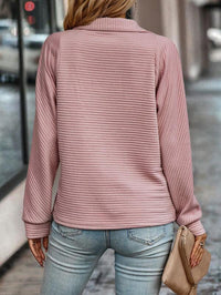 Frau in rosa Strickpullover und blauer Jeans, hält beige Clutch, Stadtstraße im Hintergrund.
