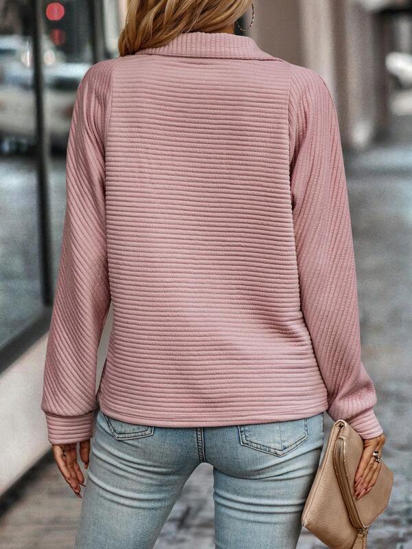 Frau in rosa Strickpullover und blauer Jeans, hält beige Clutch, Stadtstraße im Hintergrund.