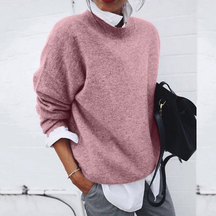 Frau in rosa Strickpullover, weißer Bluse, grauer Hose, schwarze Tasche, lässiger Stil.
