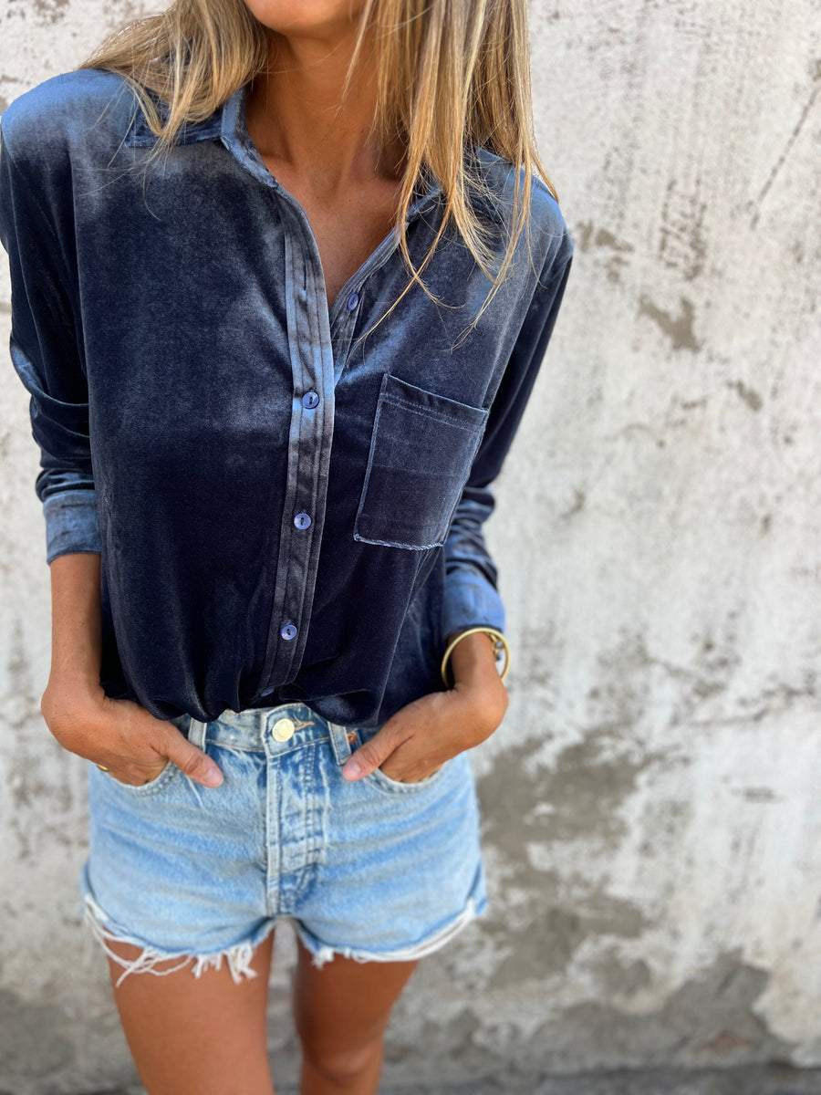 Frau in dunkelblauer Samtbluse und Jeansshorts vor Betonwand.