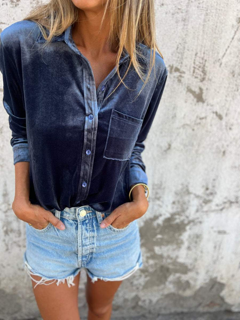 Frau in dunkelblauer Samtbluse und Jeansshorts vor Betonwand.