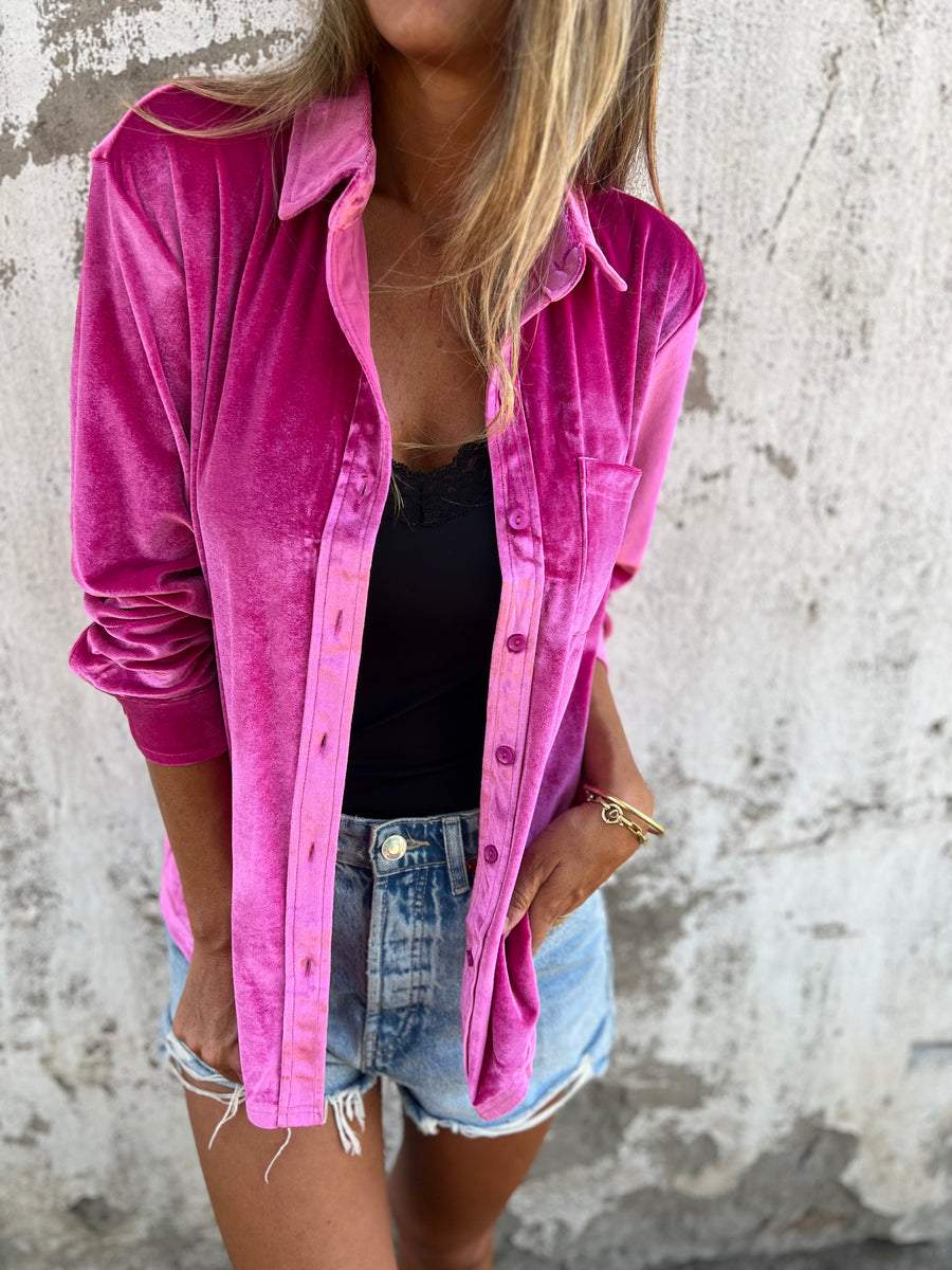 Frau in pinker Samtjacke, schwarzem Top und Jeansshorts vor Betonwand.