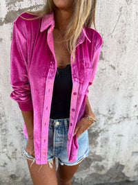 Frau in pinker Samtjacke, schwarzem Top und Jeansshorts vor Betonwand.