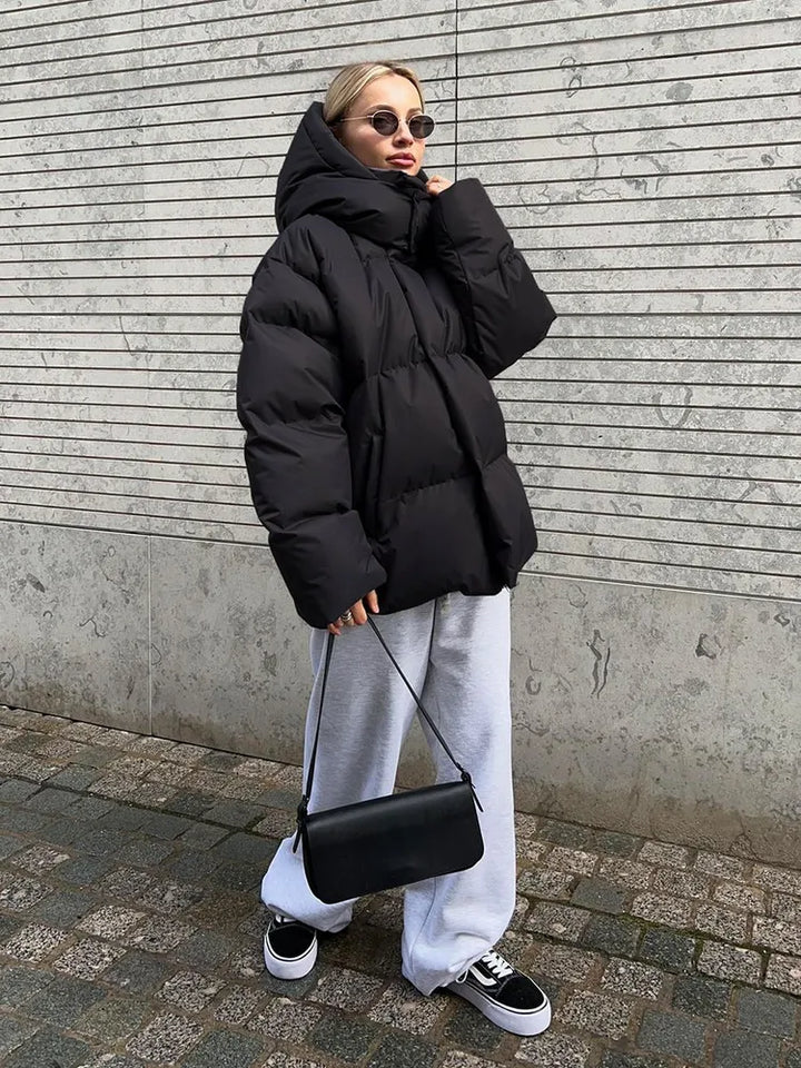 Frau in schwarzer Daunenjacke, grauer Jogginghose, Sonnenbrille, schwarze Tasche, Sneaker.