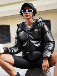 Frau in schwarzer Daunenjacke, Sonnenbrille und Mütze, hält Kaffeetasse, urbaner Hintergrund.