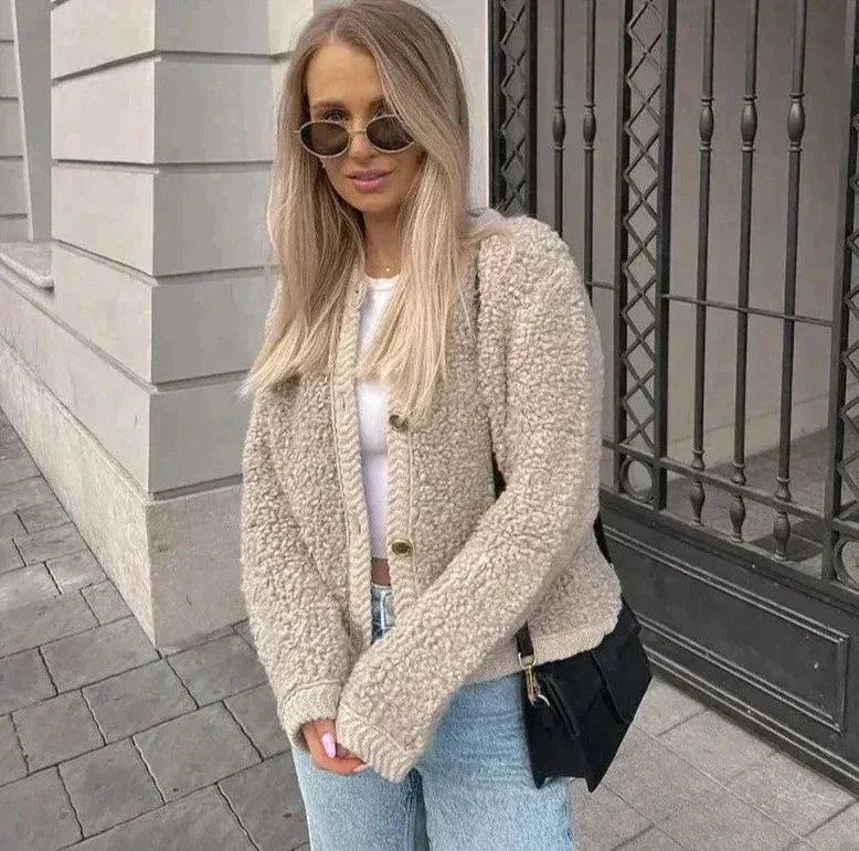 Frau in beiger Teddyjacke, blauer Jeans, Sonnenbrille, schwarze Tasche, vor Gebäude.