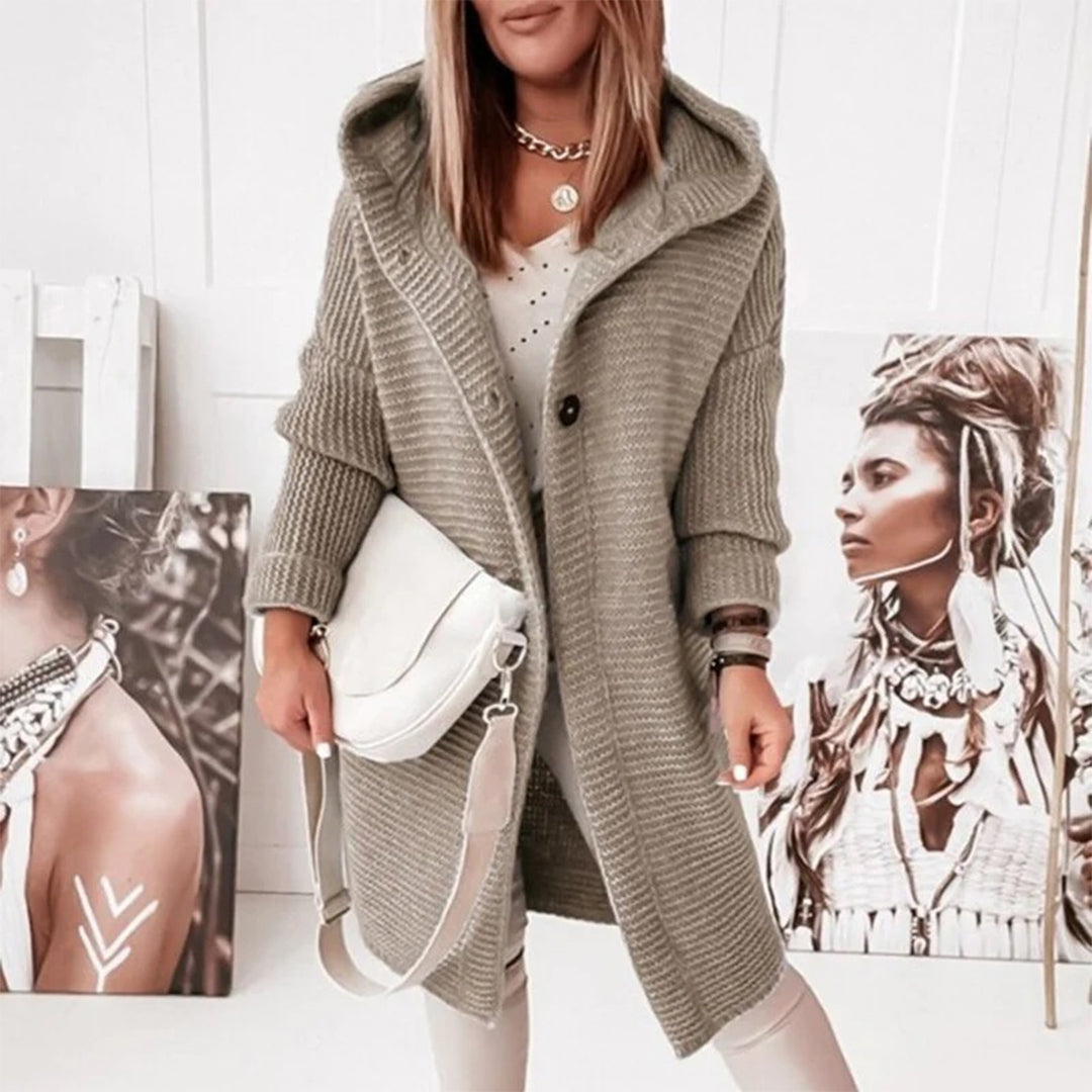 Frau in grauem Strickmantel mit Kapuze, hält weiße Umhängetasche, modisches Outfit.