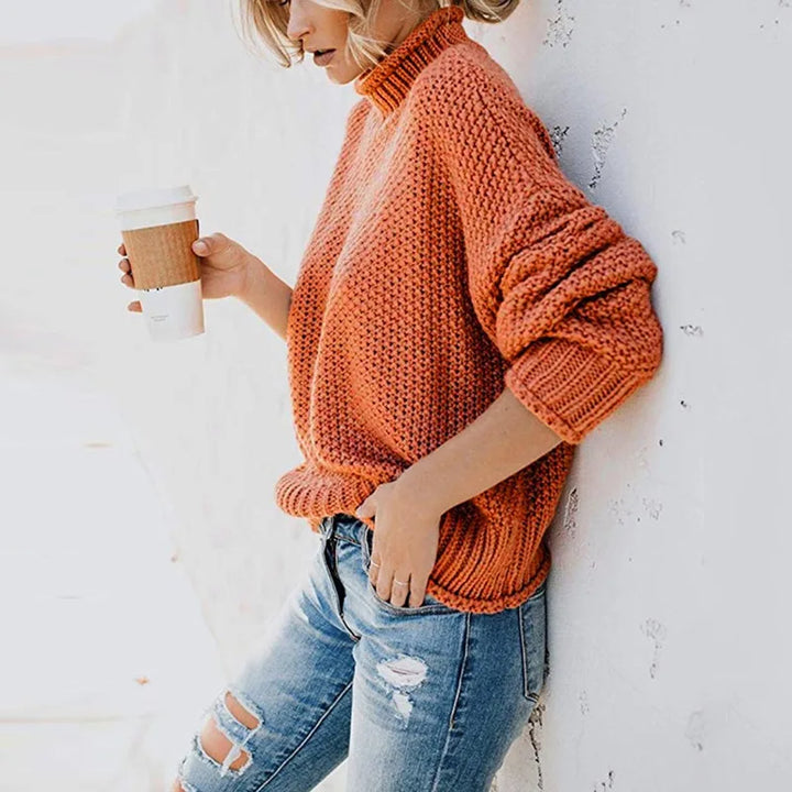 Frau in orangefarbenem Strickpullover, hält Kaffeebecher, trägt zerrissene Jeans.