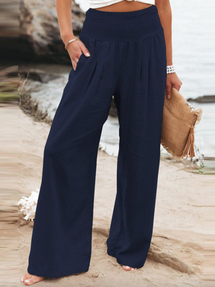 Frau in blauer, weiter Hose am Strand, hält beige Tasche, Sommermode, Damenbekleidung.
