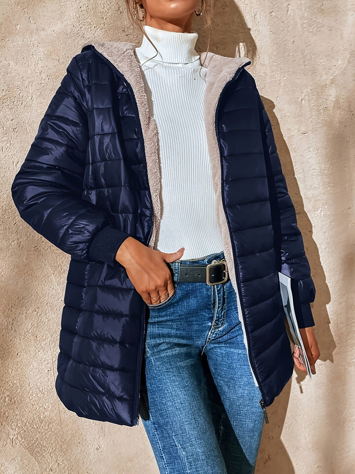 Frau in dunkelblauer Steppjacke, weißem Rollkragenpullover und Jeans vor beiger Wand.