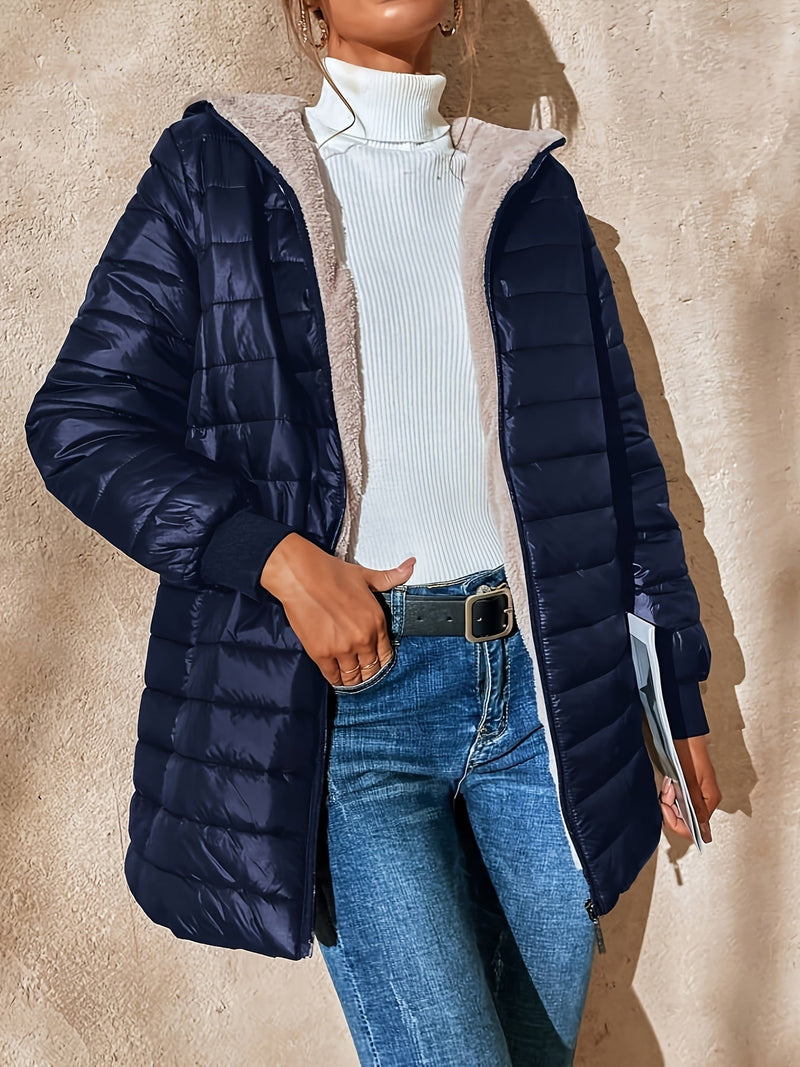 Frau in dunkelblauer Steppjacke, weißem Rollkragenpullover und Jeans vor beiger Wand.