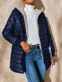 Frau in dunkelblauer Steppjacke, weißem Rollkragenpullover und Jeans vor beiger Wand.