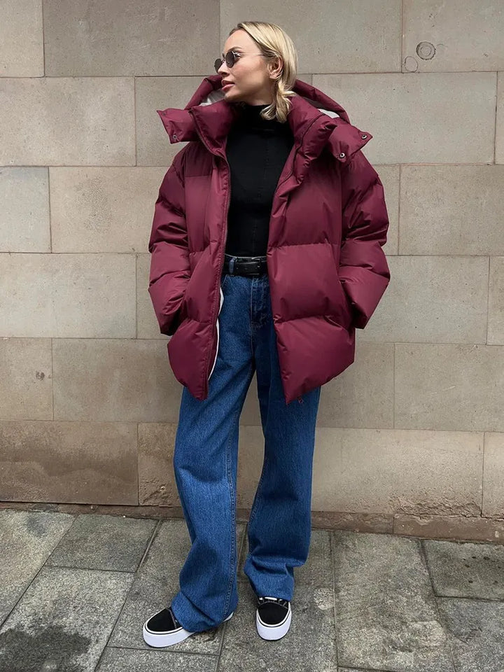 Frau in bordeauxroter Daunenjacke, schwarze Sonnenbrille, blaue Jeans, schwarze Sneaker.