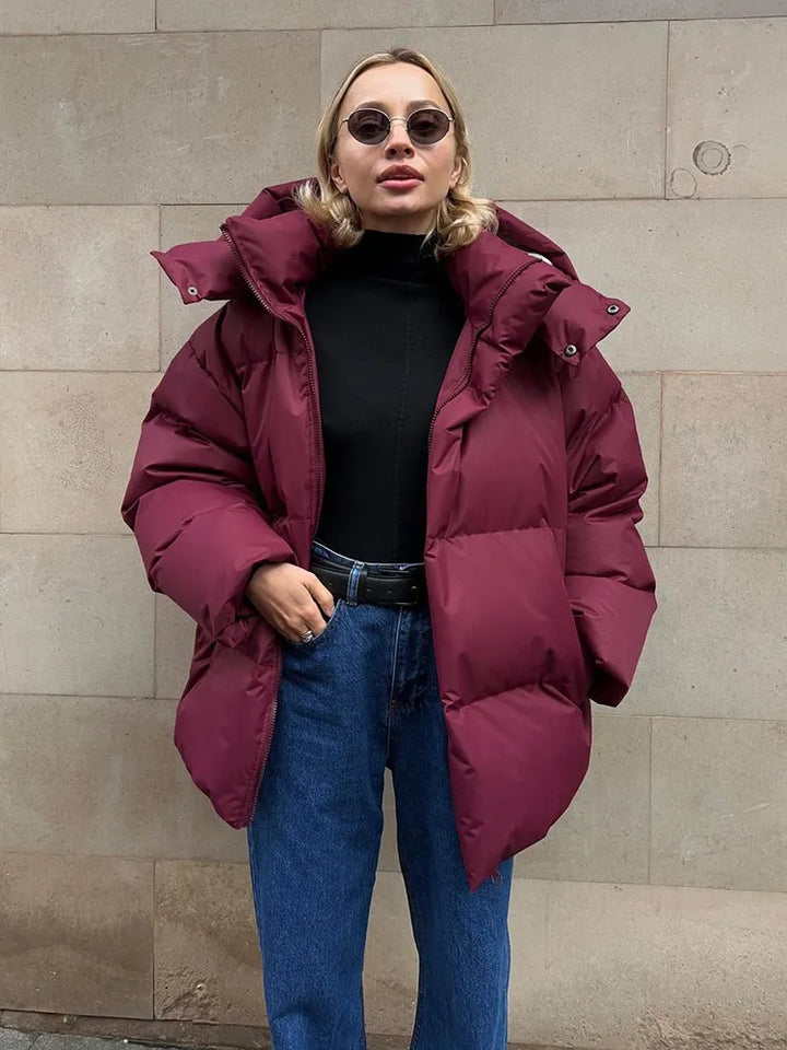 Frau in bordeauxroter Daunenjacke, schwarze Sonnenbrille, Jeans, modischer Winterlook.