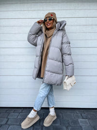 Frau in grauer Daunenjacke, Jeans, Mütze, Sonnenbrille, hält weiße Tasche, Wintermode.