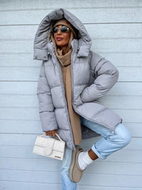 Frau in grauer Daunenjacke mit Kapuze, Sonnenbrille, beige Tasche, lässiger Winterlook.