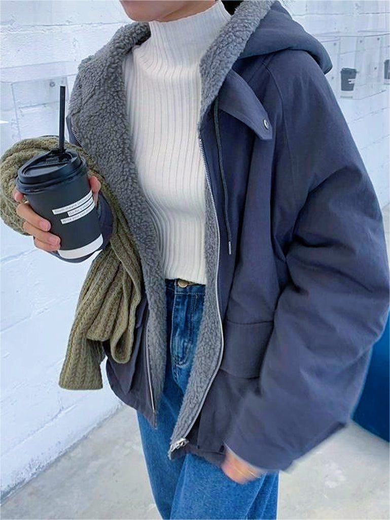Frau in dunkelblauer Winterjacke mit Kapuze, weißem Rollkragenpullover, hält Kaffeebecher.