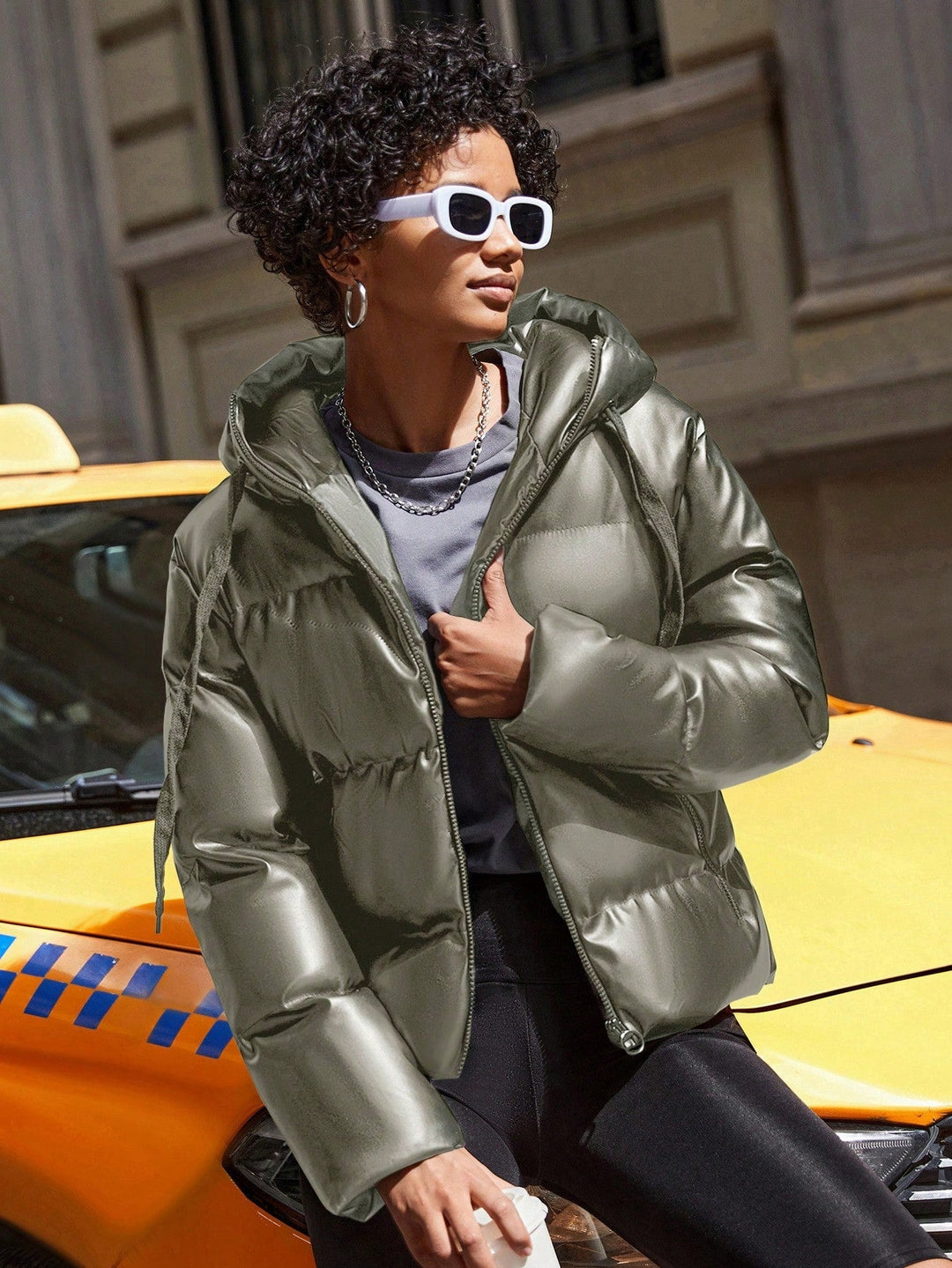 Frau mit silberner Daunenjacke und Sonnenbrille vor gelbem Taxi in der Stadt.