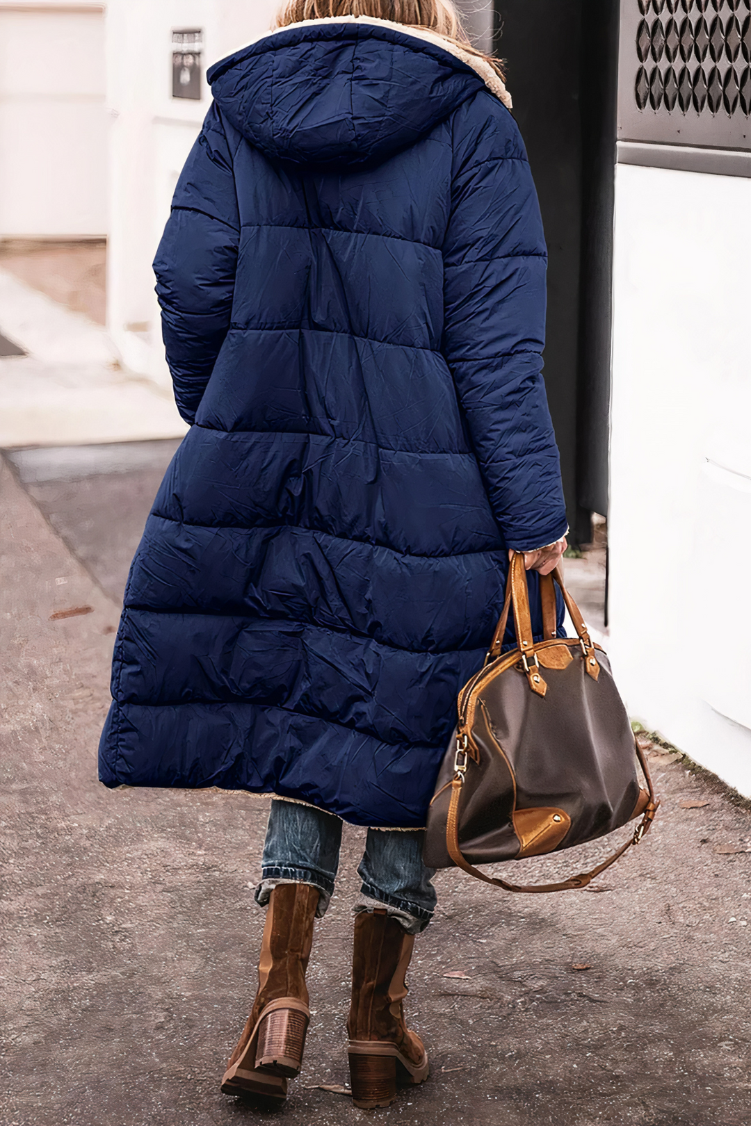 Frau in blauer Daunenjacke mit Kapuze, braune Stiefel, braune Ledertasche, Straße im Hintergrund.