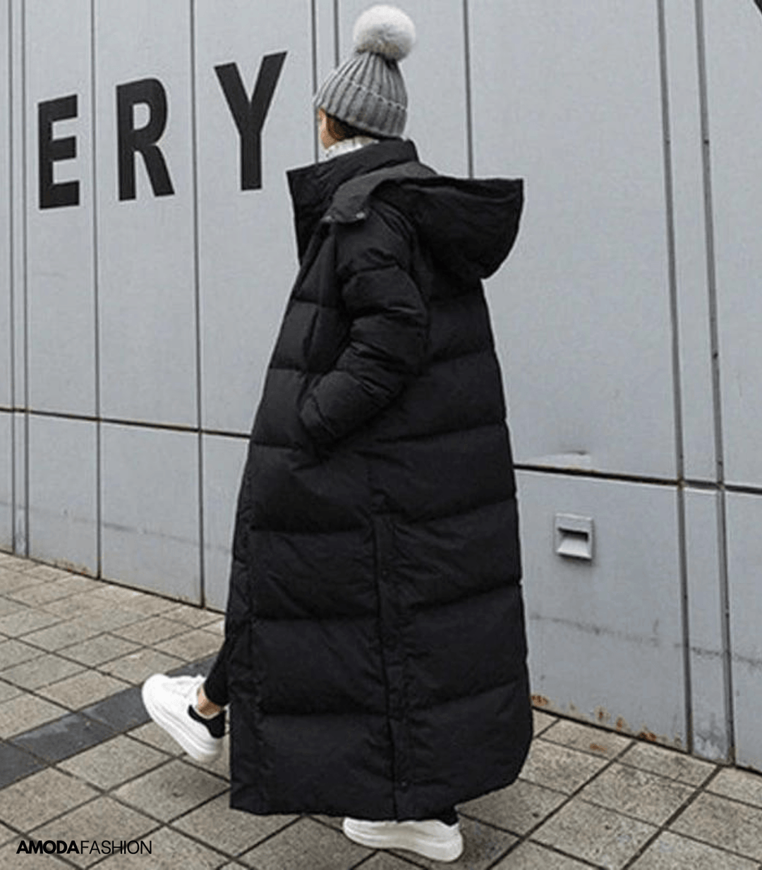 Frau im schwarzen, übergroßen Winterparka mit Kapuze, graue Mütze, weiße Sneaker, urbaner Hintergrund.