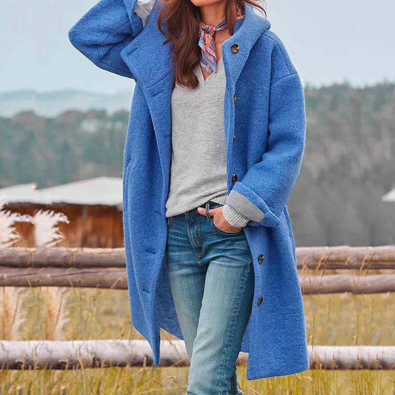 Frau in blauem Wollmantel, grauem Pullover und Jeans vor ländlicher Kulisse.